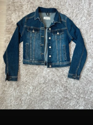 jeansjacka  - Klassisk mörkblå jeansjacka från märket Reversed med raka sömmar, silverfärgade knappar och två bröstfickor med lock. Tillverkad i denim med kontrastsömmar för en tidlös look.