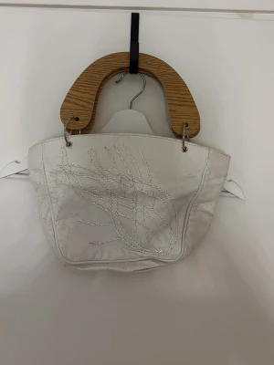 Vit handväska med trähandtag - Unik vit handväska i skinn med dekorativa sömmar på framsidan och stora handtag i trä. Väskan har en mjuk, rundad form och metallringar som fäster handtagen. Perfekt för dig som gillar statement-accessoarer.