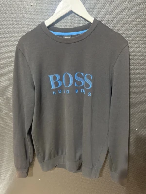 Svart sweatshirt från Hugo Boss - Snygg svart sweatshirt från Hugo Boss med blå logga tryckt på bröstet. Tröjan har rund halsringning, långa ärmar och ribbade muddar vid ärmslut och nederkant. Perfekt för en clean och stilren look.