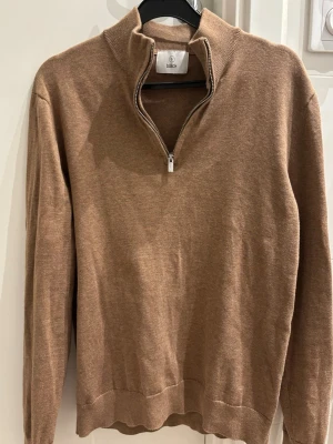 Beige stickad tröja med halvzip Bläck - Snygg beige stickad tröja från Bläck med halvzip och hög krage. Tröjan har långa ärmar och ribbade muddar vid ärmslut och nederkant. Perfekt för lager på lager och enkel att matcha med olika outfits.