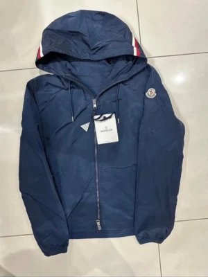 Moncler  - Snygg marinblå vindjacka från Moncler med huva och dragkedja framtill. Jackan har vita och röda detaljer på huvan samt Moncler-logga på ärmen. Tillverkad i lätt och slitstarkt material, perfekt för blåsiga dagar. Justerbar huva med dragsko och elastiska muddar. Storlek 1/S-M-X. Pris kan diskuteras vid snabbaffär. Med NFC-tag.