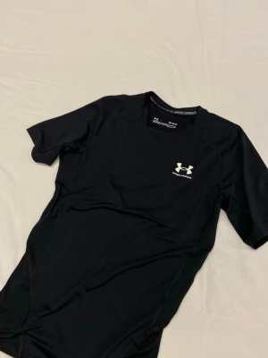 Under Armour compression shirt - Svart kortärmad träningströja från Under Armour med logga på bröstet och nacken. Tillverkad i ett stretchigt och fukttransporterande material som passar perfekt för intensiva träningspass. Snygg passform och diskreta sömmar för extra komfort.