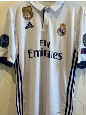 Real Madrid matchtröja Sergio Ramos - Säljer en vit Real Madrid matchtröja från Adidas med mörkblå detaljer, Champions League-märken på ärmarna och FIFA-märke på bröstet. Tryck med Sergio Ramos och nummer 4 på ryggen. Materialet är lätt och andas, perfekt för fotbollsträning eller samling.