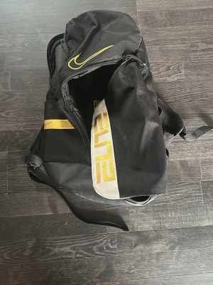 Svart Nike Elite ryggsäck - Snygg svart Nike Elite ryggsäck med guldfärgade detaljer och logga. Ryggsäcken har flera fack, justerbara axelband och en stor ELITE-text på framsidan. Tillverkad i slitstarkt material, perfekt för träning eller skola.