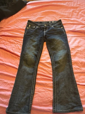 Miss me jeans - Säljer ett par mörkblå bootcut jeans i mid rise från Miss me med snygga broderade bakfickor i guld och lila. Jeansen har kontrastsömmar  och dekorativa nitar. Perfekta för dig som vill sticka ut lite extra med detaljerad design. Säljer även på vinted och pris kan alltid diskuteras det är ett dyrt märke 💕