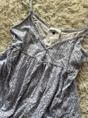 Blommig babydoll topp från Monki - Söt blå linnetopp från Monki med små vita blommor, tunna axelband och v-ringning med knappar framtill. Toppen har en luftig passform och är perfekt för varma dagar. Tillverkad i mjuk bomull som känns skön mot huden.