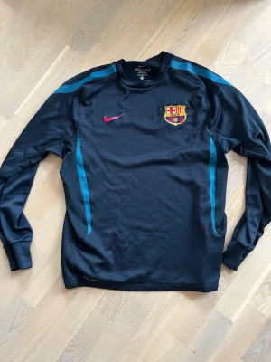 Retro 2010/2011 FC Barcelona tröja köpt i Barca  - Säljer en svart långärmad träningströja från Nike med FC Barcelona-emblem på bröstet och rosa Nike-logga. Tröjan har blå detaljer längs ärmarna och är tillverkad i Dri-FIT material som andas, perfekt för fotbollsträning eller gymmet.