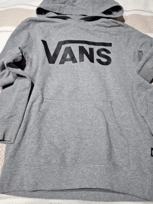 Grå hoodie från Vans med logga - Säljer en grå hoodie från Vans med stor svart logga på bröstet. Hoodien har huva och känguruficka framtill. Tillverkad i mjukt bomullsmaterial som är skönt att bära. Perfekt för dig som gillar streetwear och enkel stil. St 155