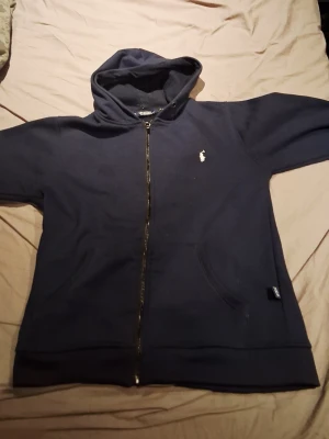 Mörkblå hoodie från Polo Ralph Lauren - Mörkblå hoodie med dragkedja från Polo Ralph Lauren. Klassisk design med huva, fickor framtill och den ikoniska vita polospelaren broderad på bröstet. Tillverkad i mjukt bomullsmaterial som är perfekt för en avslappnad stil.