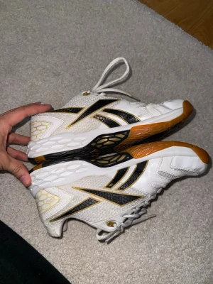 Vita Reebok inomhusskor med  - Säljer ett par vita inomhusskor från Reebok med svarta och guldiga detaljer. Perfekta för sporter inomhus som handboll eller innebandy. 