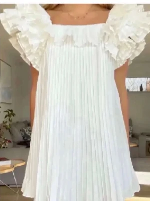 Vit plisserad klänning med volang - Supersöt vit klänning med plisserat tyg och stora volangdetaljer vid axlarna. Klänningen har en luftig passform och är ärmlös, perfekt för sommarens alla tillfällen. Ger en romantisk och trendig vibe.