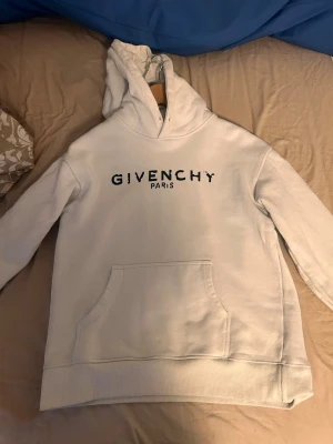 Vit hoodie från Givenchy Paris - Snygg vit hoodie från Givenchy Paris med svart tryck på bröstet. Klassisk känguruficka framtill och stor huva. Tillverkad i mjukt bomullsmaterial som känns skönt mot huden. Perfekt för dig som vill ha en stilren och exklusiv look.