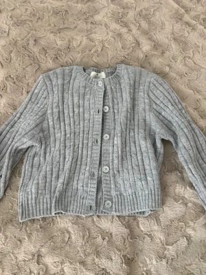 Grå kabelstickad kofta med knappar - Grå kabelstickad kofta med rund hals och vita knappar framtill. Koftan har ribbade muddar och en croppad passform som ger en trendig look. Perfekt att slänga över ett linne eller t-shirt för en chill stil.