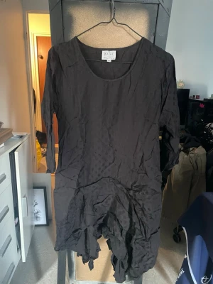 Svart klänning från Masai - Svart klänning från Masai med trekvartsärm och rund halsringning. Klänningen har asymmetrisk nederkant och lös passform, vilket ger en avslappnad och unik siluett. Tillverkad i ett lätt material som känns skönt mot huden.