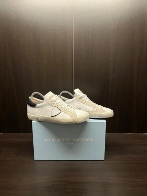 Philippe Model vita sneakers - Stilrena vita sneakers från Philippe Model med beige detaljer och svart hälparti. Skorna har en klassisk låg siluett, vita snören och en robust sula. Tillverkade i läder med mockadetaljer och ikonisk sköldlogga på sidan. Perfekta för en trendig och avslappnad look.