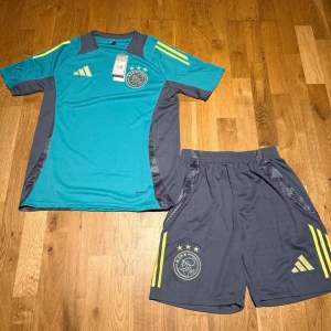 Ajax summer set turkos - Säljer ett Ajax träningsset från Adidas med en turkos t-shirt och grå shorts. Båda delar har gula detaljer och Ajax-logga med tre stjärnor. Materialet är lätt och andas, perfekt för fotboll eller träning. T-shirten har korta ärmar och rund hals.