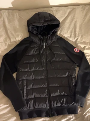 Canada goose cardigan  - Säljer en svart pufferjacka från Canada Goose med huva och dragkedja. Jackan har quiltad front och ärmar i ett mjukare material. Ikonisk Canada Goose-logga på ärmen. Perfekt för kalla vinterdagar och riktigt snygg att bära.
