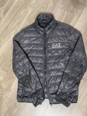 Svart vindjacka från EA7 Emporio Armani - Snygg svart vindjacka från EA7 Emporio Armani med quiltad design och diskret logga på bröstet. Jackan har dragkedja framtill, hög krage och är tillverkad i lätt, glansigt syntetmaterial. Perfekt för dig som vill ha en stilren och sportig look.