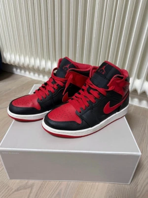Air Jordan 1 Mid röd/svart - Säljer ett par Air Jordan 1 Mid sneakers i rött och svart läder med vita sulor och röda skosnören. Klassisk Jumpman-logga på plösen och ikonisk Swoosh på sidan. Perfekta för dig som gillar streetwear och vill sticka ut med en tidlös modell.