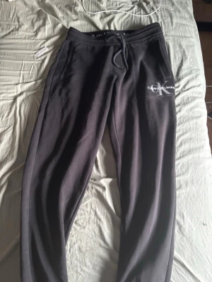 Svarta mjukisbyxor från Calvin Klein - Svarta mjukisbyxor från Calvin Klein Jeans med snöre i midjan och fickor på sidorna. Broderad CK-logga på bakfickan och tryckt logga framtill. Sköna och stilrena, perfekta för chill eller streetwear. Mjukt material, troligtvis bomullsblandning. Storlek S andvänada typ 3 gånger 