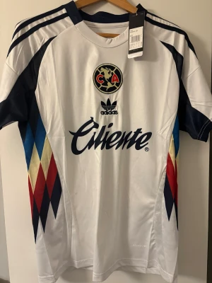 Club América Adidas matchtröja  - Snygg vit matchtröja från Adidas för Club América med klubbmärke på bröstet och Caliente-logga. Tröjan har mörkblå detaljer, färgglada diamantmönster i rött, blått och gult på sidorna samt klassiska Adidas-ränder på axlarna. Tillverkad i lätt funktionsmaterial.