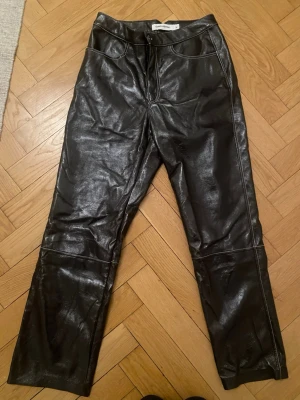 Svarta coated jeans från Gina Tricot - Bruna byxor från Carin Wester  från med raka ben och hög midja. Byxorna har en glansig finish som ger en cool läderlook och synliga sömmar längs benen. Går till ankeln. Se mått på bild. 