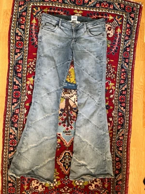 Ljusblå lågmidjade bootcut jeans från BDG - Säljer ett par ljusblå bootcut jeans från BDG med diagonal sömdetaljer och klassiska fem fickor. Jeansen har en bred utsvängning nedtill och är tillverkade i denim. Perfekt för dig som gillar retrostil och vill sticka ut.