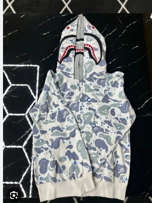 BAPE haj-hoodie med camomönster - Säljer en ikonisk BAPE zip hoodie med hajmotiv på huvan och gråblått camomönster över hela plagget. Dragkedja framtill, ribbade muddar och fickor på sidorna. Perfekt för dig som gillar streetwear och vill sticka ut med en unik design.