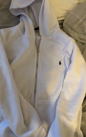 Vit hoodie från Polo Ralph Lauren - Säljer en vit hoodie från Polo Ralph Lauren med dragkedja framtill och klassisk huva. Tröjan har det ikoniska lilla Polo-logotypen broderad på bröstet i mörkblått. Tillverkad i mjukt och bekvämt material, perfekt för en avslappnad stil.