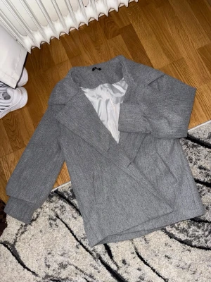 Grå kappa med bred krage - Snygg grå kappa med bred krage och rak passform. Kappan har två sidofickor och är helfodrad med ljusgrått innerfoder. I bra skick men är nopprig och insidan av fodret har också ett hål i sig, inget som påverkar yttre utseendet på kappan som man kan se på bild 4. Hör av er för fler bilder/ frågor!!