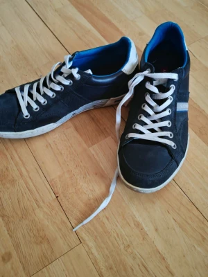 Björn Borg sneakers - Snygga svarta sneakers från Björn Borg med vita sulor och snören, blå insida och vita detaljer på hälen. Skorna har en klassisk låg profil och är tillverkade i en mix av mocka och textil. Perfekta för en avslappnad streetstyle-look.