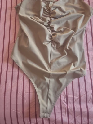 Beige body med rynk framtill - Snygg beige body med rynkdetalj framtill och tryckknappar i grenen. Tillverkad i mjukt, stretchigt material som sitter skönt mot kroppen. Perfekt att styla till jeans eller kjol för en clean look.