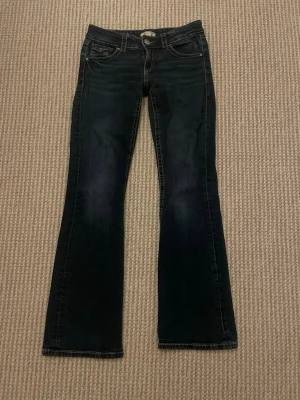 Mörkblå bootcut jeans med fickor - Snygga mörkblå bootcut jeans med klassiska fem fickor och kontrastsömmar. Jeansen har låg midja och lätt utsvängda ben. Tillverkade i denim med en skön stretch för extra komfort. Perfekta för en avslappnad och trendig look.