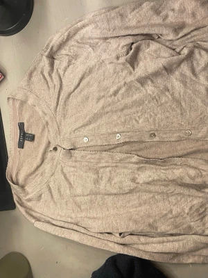 Beige cardigan från Esprit - Ljusbeige tunn cardigan från Esprit med rund halsringning och knäppning framtill. Tröjan har långa ärmar och smala ribbade muddar. Perfekt att slänga över en topp för en enkel och clean look.