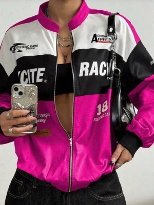 Färgstark racing bomberjacka storlek M - Säljer en riktigt snygg bomberjacka i stark rosa, svart och vit med racing-inspirerade tryck och detaljer. Jackan har dragkedja framtill, ribbade muddar och coola texter som 'RACITE', 'CHICAGO' och 'EXCITING GAME'. Perfekt statement-plagg för dig som vill sticka ut.