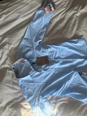 Ljusblå skjorta från Burberry - Säljer en ljusblå skjorta från Burberry helt oanvänd. Skjortan har knappar framtill och är långärmad. Perfekt för sommaren.