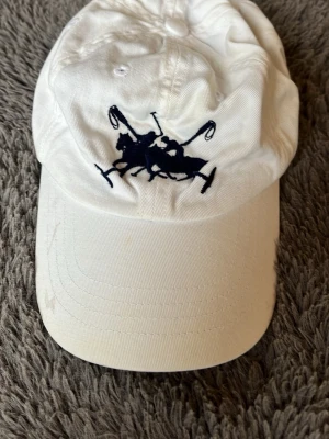 Vit keps från Polo Ralph Lauren - Snygg vit keps från Polo Ralph Lauren med mörkblå broderad polospelare framtill och texten 'POLO' broderad på sidan. Klassisk böjd skärm och justerbart spänne bak. Tillverkad i bomull för en bekväm och luftig känsla.