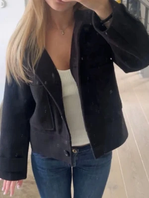 Svart ullkappa från Kappahl - Stilren svart kappa från Kappahl i ullmix med rak, boxig passform och stora utanpåfickor. Kappan har bred krage, knäppning framtill och är midjekort. Perfekt för våren. Inte särskilt använd och i gott skick