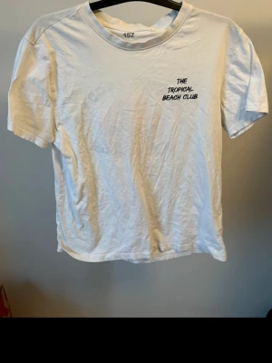 Vit t-shirt The Tropical Beach Club - Vit t-shirt från 157 med tryck 'The Tropical Beach Club' fram och stort tryck med sol och text bak. Klassisk rund hals och korta ärmar. Perfekt för en avslappnad och somrig stil.