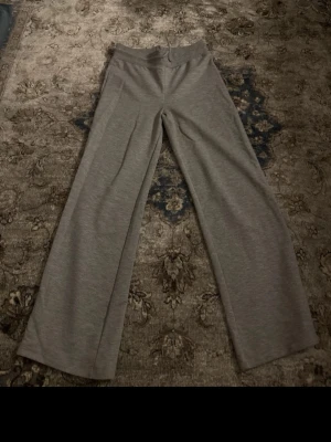 Grå mjukisbyxor med dragsko - Säljer ett par grå mjukisbyxor med bred resår i midjan och dragsko för skön passform. Byxorna har raka ben och är gjorda i mjukt, stretchigt material som passar perfekt för chill dagar. Enkla och stilrena utan tryck eller detaljer.