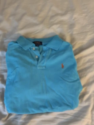 Blå pikétröja från Polo Ralph Lauren - Säljer en klassisk ljusblå pikétröja från Polo Ralph Lauren med den ikoniska broderade loggan i orange på bröstet. Tröjan har krage, knappar framtill och korta ärmar. Perfekt till en avslappnad men ändå stilren look.