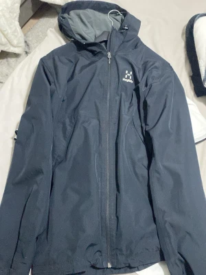 Svart vindjacka från Haglöfs - Snygg svart vindjacka från Haglöfs med huva och hel dragkedja framtill. Jackan har minimalistisk design, diskret logga på bröstet och är tillverkad i ett lätt, vindtätt material. Perfekt för blåsiga dagar och outdoor-äventyr.