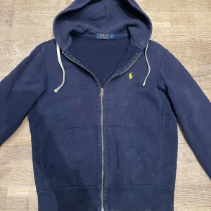 Mörkblå hoodie från Polo Ralph Lauren - Mörkblå hoodie med dragkedja från Polo Ralph Lauren. Klassisk design med huva och vita dragsnören, kängurufickor framtill och den ikoniska gula logotypen broderad på bröstet. Tillverkad i mjuk bomullsblandning som känns skön mot huden. Storlek S! Pris kan diskuteras