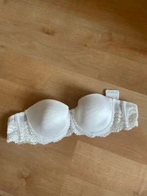 Vit axelbandslös bh med spets - Säljer en vit axelbandslös bh med vadderade kupor och fina spetsdetaljer längs kanterna. Bh:n har bygel och bred rygg med hakknäppning. Perfekt under toppar och klänningar där du vill slippa axelband.