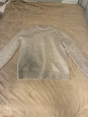 Ljusgrå stickad tröja unisex - En enkel och stilren ljusgrå stickad tröja med rund hals och ribbade muddar vid ärmslut och nederkant. Tröjan har en rak passform och är perfekt för lager-på-lager. Materialet ser ut att vara mjukt och bekvämt, troligen ull- eller bomullsblandning. Köpt på en marknad.