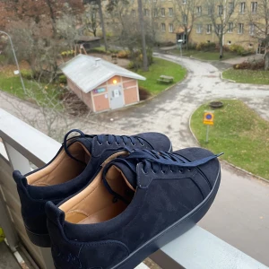 Mörkblå sneakers från Christian Louboutin - Säljer ett par mörkblå sneakers från Christian Louboutin i mocka med klassisk röd sula och snygg broderad logga på hälen. Skorna har rund tå, platt sula och snörning. Insidan är ljusbeige och de har en stilren, minimalistisk look som sticker ut med den ikoniska röda undersidan. Storlek 46 men passar för storlek 45, och oanvända, nya. Vid frågor bara att skriva!