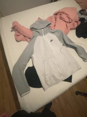 Nike tech (old season)  - Snygg grå hoodie från Nike med dragkedja framtill och två stora fickor. Tröjan har ljusgrå kropp och mörkare grå ärmar samt en klassisk huva. Nike-loggan är broderad på bröstet. Perfekt för en avslappnad och sportig stil.