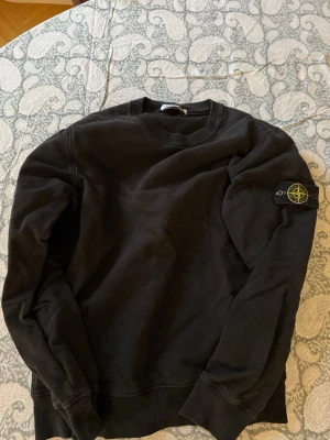 Svart bomulls tröja Stone Island - Svart bomulls tröja från Stone Island med rund hals och patch på vänster ärm. Tröjan har ribbade muddar vid ärmslut och nederkant. 