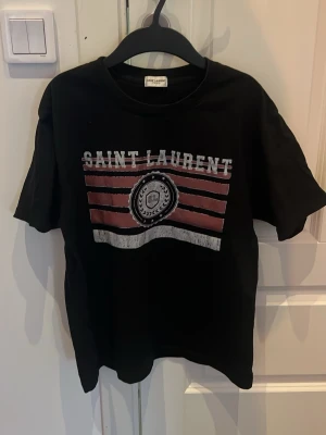 Svart Saint Laurent t-shirt - Snygg svart t-shirt från Saint Laurent med tryck i vitt och rött framtill. Klassisk rund hals och korta ärmar. Perfekt till jeans eller shorts för en cool och avslappnad look.