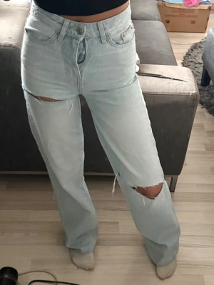Ljusblå ripped jeans med raka ben - Säljer ett par ljusblå jeans med raka ben och stora slitningar vid knäna. Jeansen har klassisk femficksmodell, hög midja och är tillverkade i denim. Perfekta för en avslappnad och trendig stil. De är aldrig använda så nya fast utan prislapp!